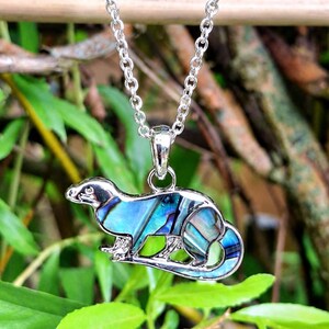 Abalone shell otter necklace hanging outdoors amongst natural greenery, featuring an iridescent paua shell otter pendant on a trace chain.