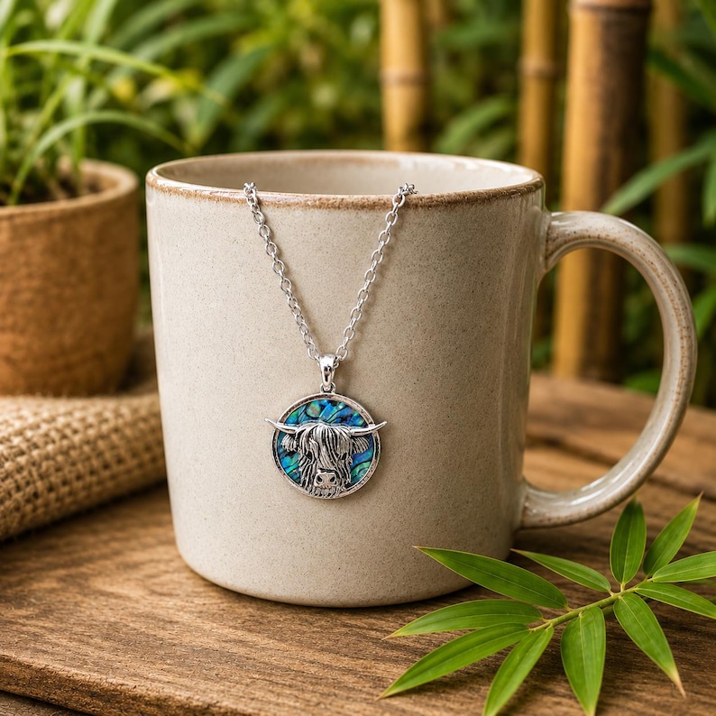 Highland cow abalone shell necklace draped over a rustic ceramic mug, featuring a silver-tone cow pendant with colourful paua shell inlay on a trace chain.