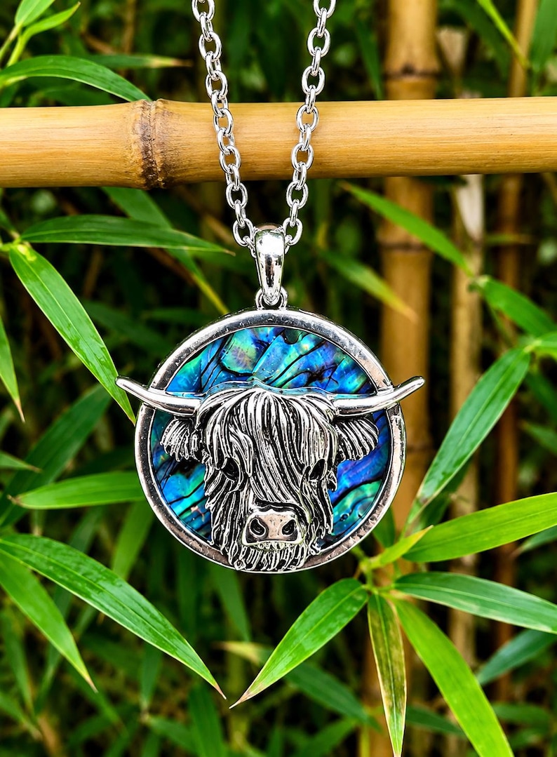 A silver-toned pendant necklace featuring a Highland cow design. The pendant has a circular frame with a blue and green iridescent background. The cow's head is detailed in silver, with horns extending outward. The necklace chain is also silver.