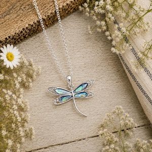 Silver dragonfly necklace with abalone shell inlay displayed on a rustic wooden surface surrounded by delicate white flowers.