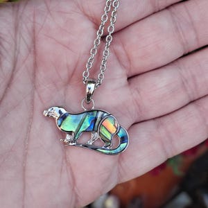 Puede incluir: Collar con colgante de nutria plateado. El colgante de nutria presenta incrustaciones iridiscentes de concha de abul&oacute;n en tonos azules, verdes y naranjas. El collar tiene una cadena delicada y un cierre de mosquet&oacute;n.