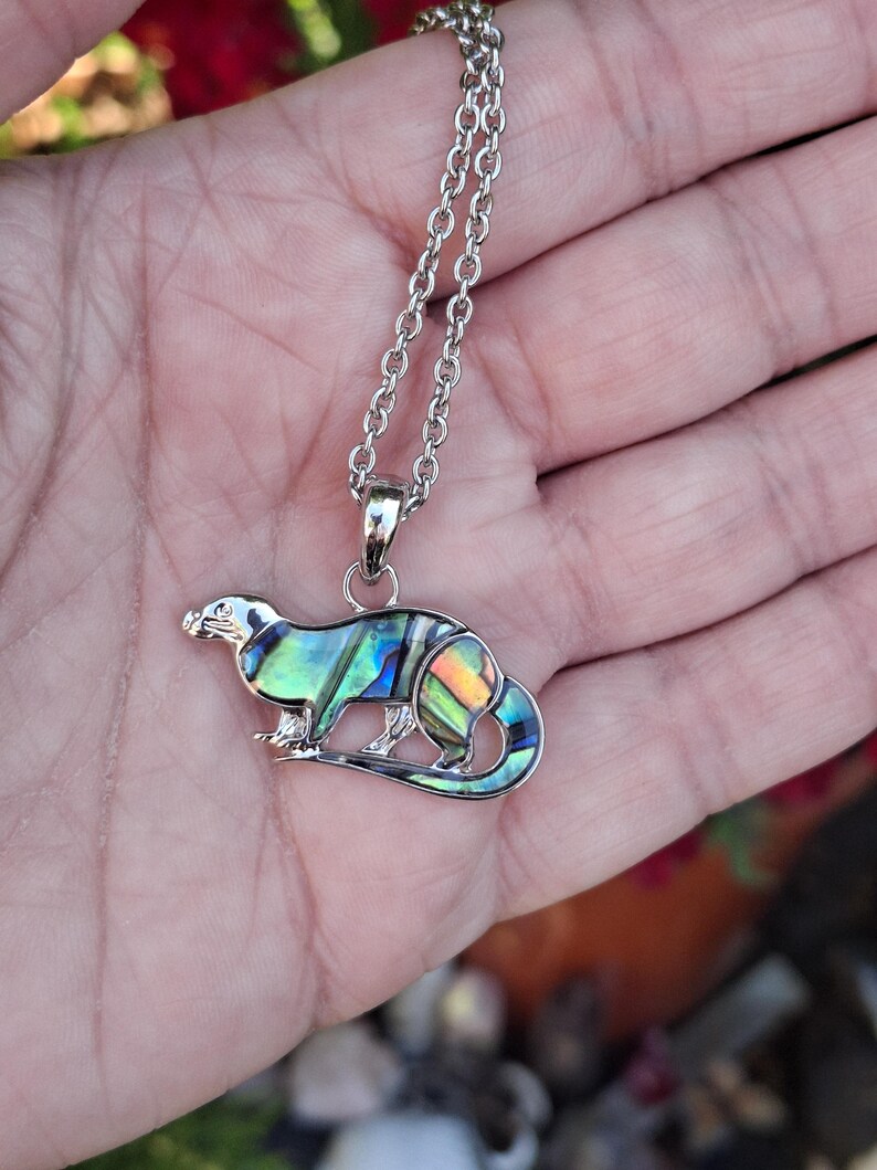 Puede incluir: Collar con colgante de nutria plateado. El colgante de nutria presenta incrustaciones iridiscentes de concha de abul&oacute;n en tonos azules, verdes y naranjas. El collar tiene una cadena delicada y un cierre de mosquet&oacute;n.