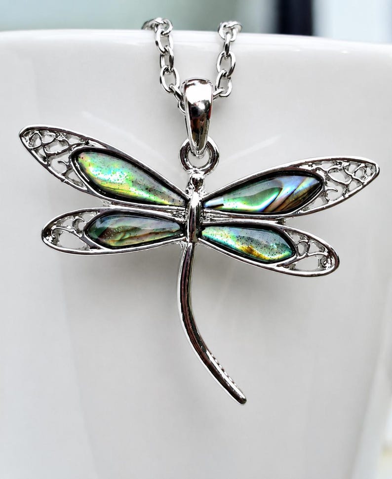 Abalone shell dragonfly necklace displayed on a white mug, highlighting the iridescent blue and green shell wings.