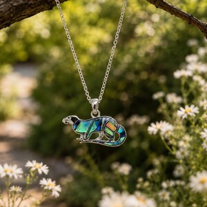 Otter pendant necklace hanging from a rustic tree branch surrounded by wildflowers, showcasing colourful abalone shell detail.