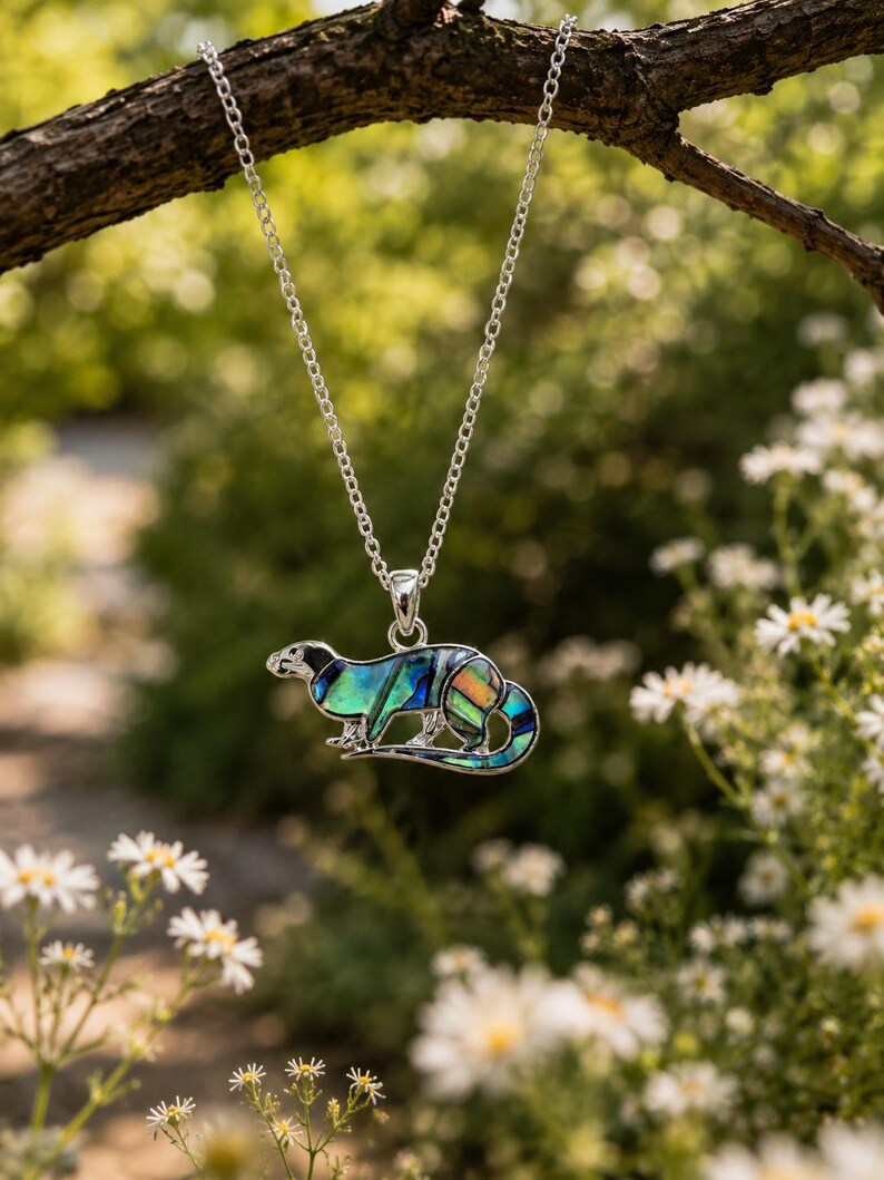 Otter pendant necklace hanging from a rustic tree branch surrounded by wildflowers, showcasing colourful abalone shell detail.