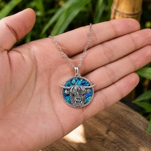 Highland cow abalone shell necklace displayed in a hand, showing the silver-tone cow pendant with colourful paua shell inlay on a trace chain.