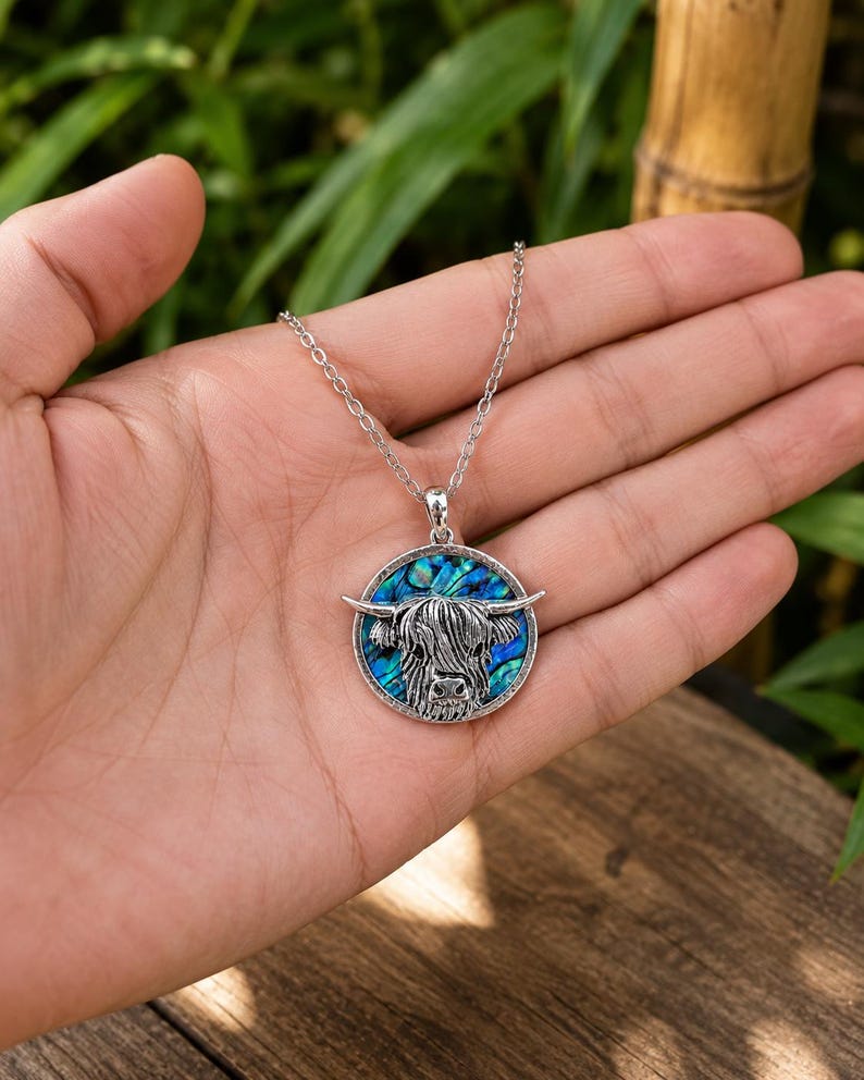 Highland cow abalone shell necklace displayed in a hand, showing the silver-tone cow pendant with colourful paua shell inlay on a trace chain.