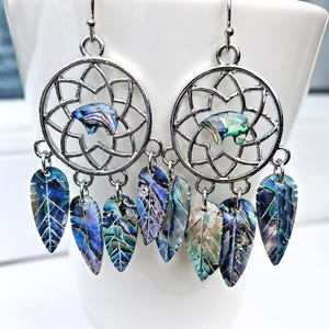 Könnte beinhalten: Silberfarbene Ohrringe mit einem Traumfänger-Design. Jeder Ohrring hat einen kreisförmigen Rahmen mit einem farbenfrohen Abalone-Muschel-Akzent und baumelnden blattförmigen Anhängern in Blau-, Grün- und Lilatönen.