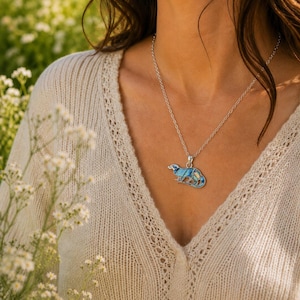Abalone shell otter necklace worn on a silver-tone trace chain, shown against a cream knit top in a natural outdoor setting. Ai model