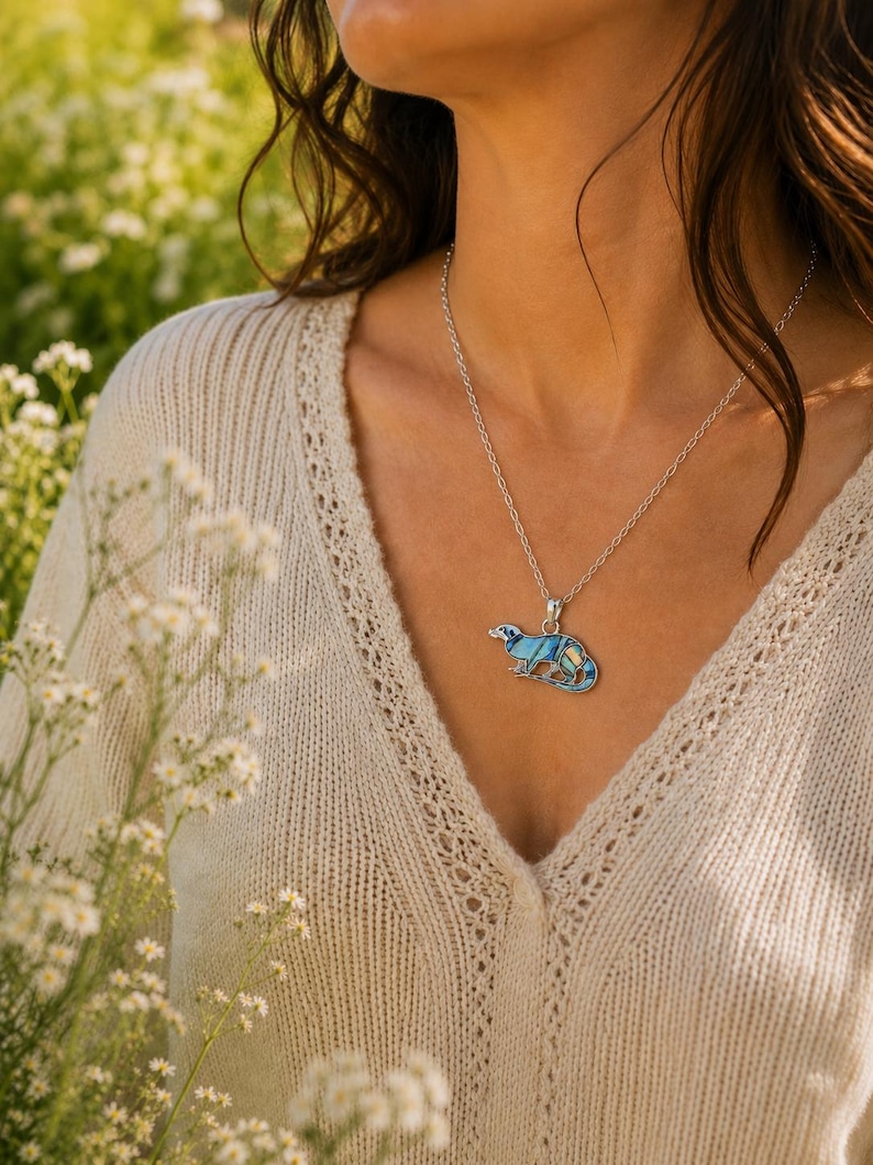 Abalone shell otter necklace worn on a silver-tone trace chain, shown against a cream knit top in a natural outdoor setting. Ai model