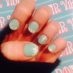 May include: A hand with light blue nail polish with white daisy accents on four nails. The thumb nail has silver glitter polish.