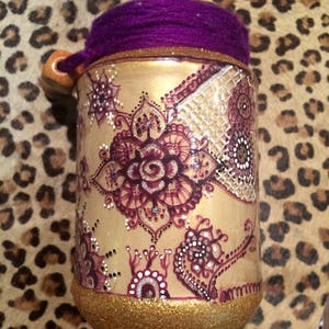 May include: A gold jar with a purple lid and intricate henna-style designs in shades of brown and white. The jar is decorated with glitter.