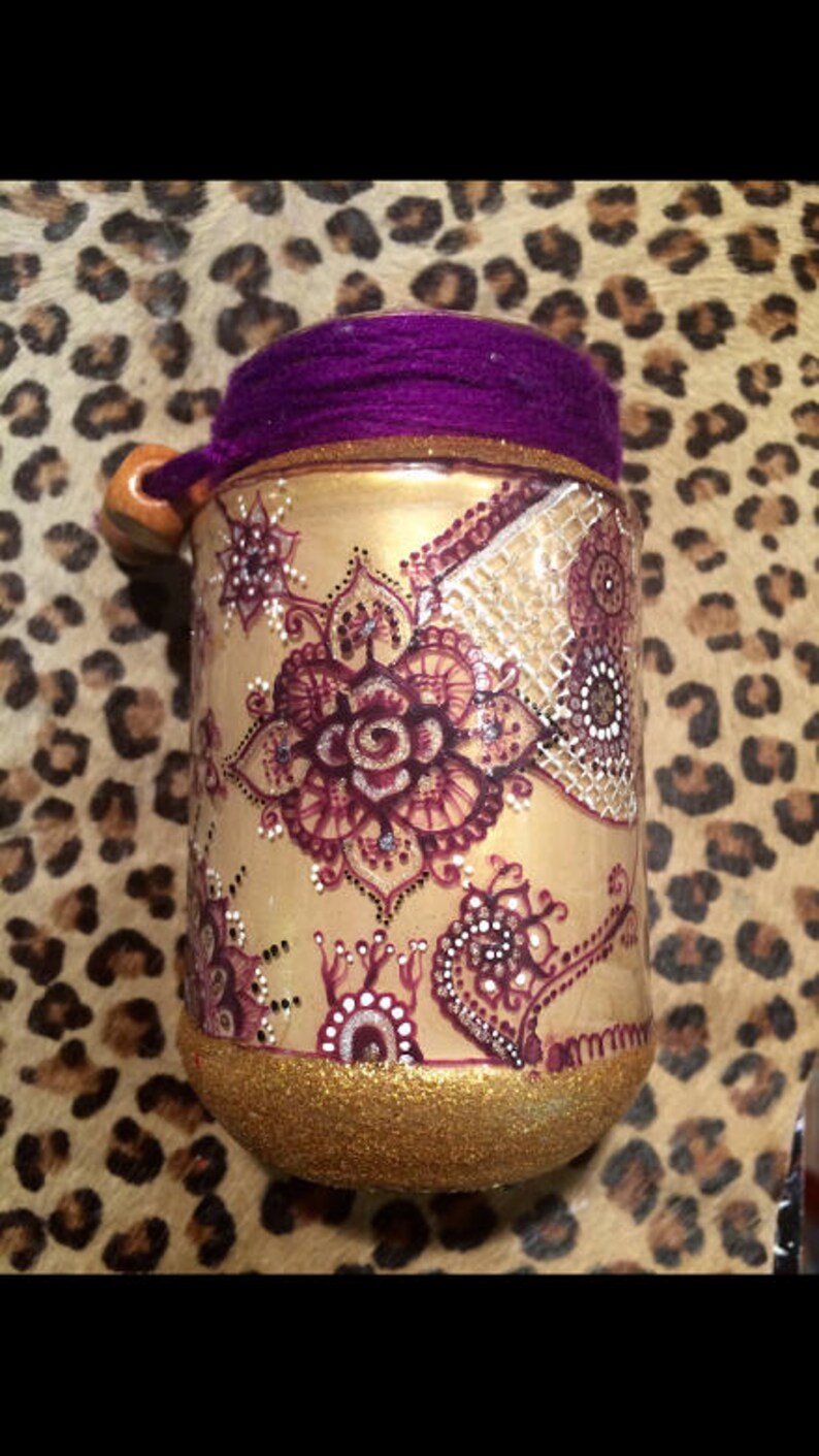May include: A gold jar with a purple lid and intricate henna-style designs in shades of brown and white. The jar is decorated with glitter.