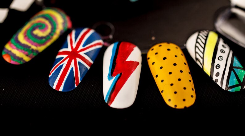 May include: Six painted fingernails with different designs. The designs include a swirl, a Union Jack, a lightning bolt, polka dots, a geometric pattern, and a striped pattern.
