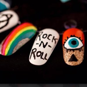 May include: Close-up of six painted fingernails with different designs. The designs include a rainbow, a black and white symbol, a white nail with the words "Rock-N-Roll" written in black, a red nail with a blue eye, and a black nail with a white triangle.