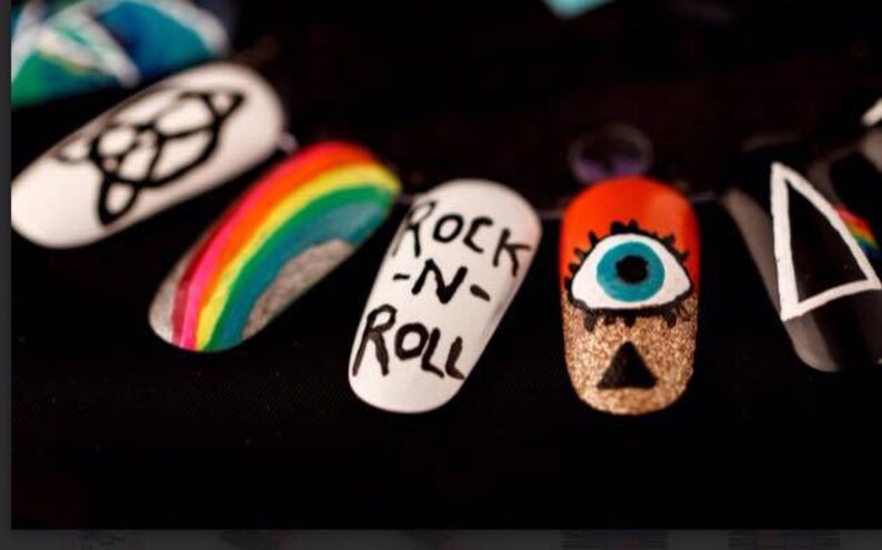 May include: Close-up of six painted fingernails with different designs. The designs include a rainbow, a black and white symbol, a white nail with the words "Rock-N-Roll" written in black, a red nail with a blue eye, and a black nail with a white triangle.
