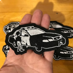 Può includere: Tre toppe ricamate in bianco e nero che rappresentano un'auto della polizia con una persona in piedi accanto.