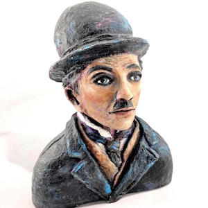 Pode incluir: Busto pintado à mão de Charlie Chaplin, com chapéu coco e fato pretos. A escultura tem um rosto detalhado com olhos azuis, bigode e expressão neutra. O busto está sobre um fundo branco.