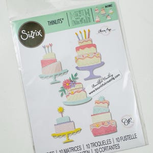May include: A Sizzix Thinlits die set in a clear plastic package containing 10 metal dies for cutting paper shapes. The packaging displays five colorful, layered cake designs with candles and floral accents, along with the text Sizzix, Thinlits, and 10 DIES.