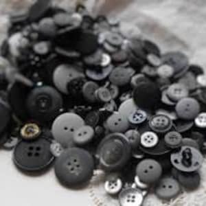 May include: A large collection of assorted sewing buttons in various shades of black, charcoal, and gray. The buttons vary in size, shape, and texture, featuring different numbers of holes and surface finishes, creating a dense, layered pile.