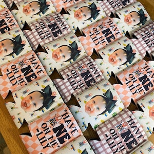 May include: A set of 12 white and pink can coolers with a checkered pattern and a baby's face. The can coolers have the text "Have One On Me - Lily" printed on them.