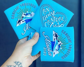 May include: A hand holds three light blue can coolers. The coolers have white text and graphics. One says "Cruisin' Together Alaska 2023". Another says "On Cruise Control". The third has a ship graphic and the words "Cruisin' Together".