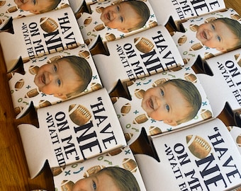 May include: A collection of white foam can coolers featuring a repeating pattern of brown footballs and green X and O symbols. Each cooler displays a photograph of a smiling baby and the black text "HAVE ONE ON ME" followed by "-WYATT REID".