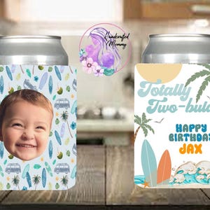 May include: Two can coolers with a beach theme. One features a smiling baby's face on a light blue background with surfboards, palm trees, and vans. The other says "Totally Two-bular" with "Happy Birthday Jax" on a beach scene.