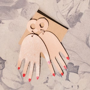 Op de afbeelding: Een paar roze handvormige oorbellen met rode nagellakaccenten. De oorbellen zijn gemaakt van hout en hebben een eenvoudig, modern ontwerp.