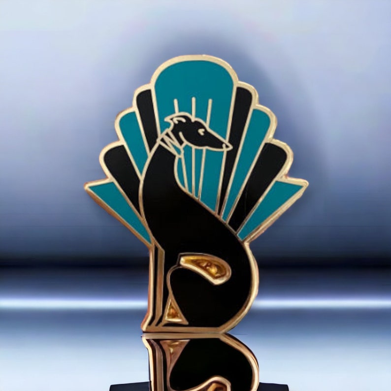 Pode incluir: Um alfinete de metal dourado com uma silhueta preta de um galgo sentado em frente a um design de leque art d&eacute;co em azul turquesa e preto.