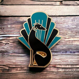 Pode incluir: Um broche de esmalte preto e dourado com um c&atilde;o galgo estilizado sentado em frente a um design de leque art d&eacute;co com detalhes em azul turquesa e preto.