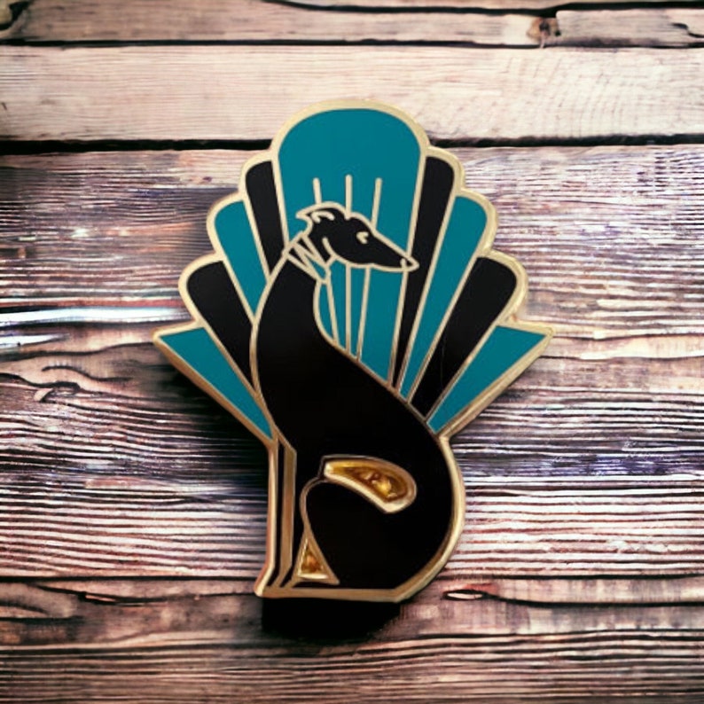 Pode incluir: Um broche de esmalte preto e dourado com um c&atilde;o galgo estilizado sentado em frente a um design de leque art d&eacute;co com detalhes em azul turquesa e preto.
