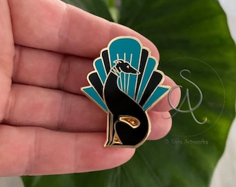 May include: Gold-tone enamel pin featuring a black silhouette of a dog sitting in front of a teal and black art deco fan. The pin reads "SASS ARTWORKS" at the bottom.