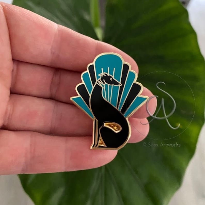 Pode incluir: Pin de esmalte dourado com uma silhueta preta de um c&atilde;o sentado em frente a um leque art d&eacute;co azul-turquesa e preto. O pin tem a inscri&ccedil;&atilde;o "SASS ARTWORKS" na parte inferior.