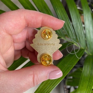 Pode incluir: Pin de metal dourado com um design detalhado de um buqu&ecirc; de flores. A parte de tr&aacute;s do pin tem dois fechos de metal para prender na roupa. O pin tem o texto "Sass Artworks" na parte de tr&aacute;s.