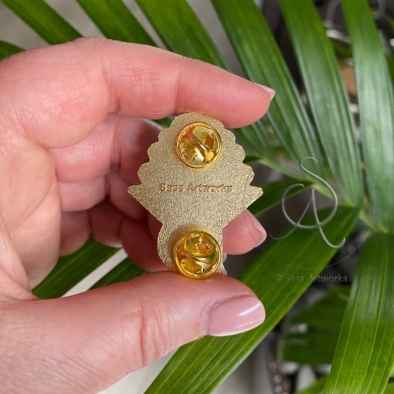 Pode incluir: Pin de metal dourado com um design detalhado de um buqu&ecirc; de flores. A parte de tr&aacute;s do pin tem dois fechos de metal para prender na roupa. O pin tem o texto "Sass Artworks" na parte de tr&aacute;s.