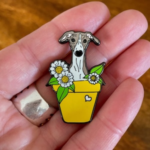 May include: A yellow enamel pin featuring a grey and white whippet dog in a yellow flower pot with white daisies and green leaves. The pot has a small heart on the side. The pin is held in a hand.