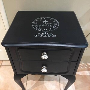 Op de afbeelding: Zwarte, gelakte houten nachtkastje met twee laden en cabriolepoten. De bovenste lade heeft een zilveren knop met Romeinse cijfers. De onderste lade heeft een zilveren knop met een bloemmotief. De bovenkant van het nachtkastje heeft een sjabloon van een wijzerplaat met het woord "PARIS" in het midden.