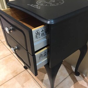 Op de afbeelding: Zwarte nachtkastje met twee laden, zilveren beslag en een stenciled klokontwerp op de bovenkant. De laden zijn open, waardoor een licht houten interieur met een zilveren stenciled ontwerp zichtbaar is.