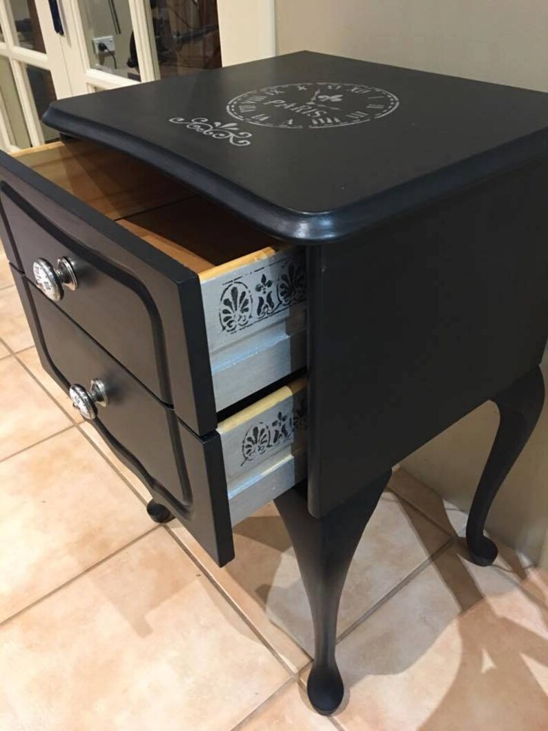 Op de afbeelding: Zwarte nachtkastje met twee laden, zilveren beslag en een stenciled klokontwerp op de bovenkant. De laden zijn open, waardoor een licht houten interieur met een zilveren stenciled ontwerp zichtbaar is.