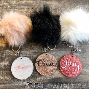 May include: Three personalized keychains with fluffy pom poms in pink, black, and white. Each keychain features a round, glitter-covered charm with a name written in cursive:  "Athena", "Olivia", and "Jenny".
