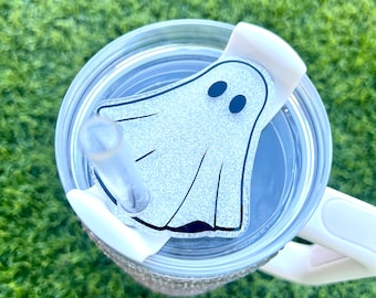 May include: A clear tumbler with a white handle and lid. The lid features a glittery ghost-shaped design with black eyes. A clear straw is inserted into the tumbler. The tumbler is designed for cold beverages.