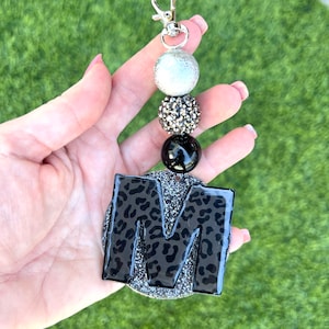 May include: A black and silver keychain with a large letter "M" charm. The charm is covered in a black and white leopard print and has a silver glitter background. The keychain also features three beads: a silver bead, a black bead with silver glitter, and a black bead.
