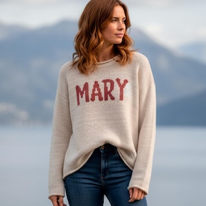 Puede incluir: Un suéter beige con el nombre "MARY" en letras rojas. El suéter se combina con unos vaqueros azules. El fondo presenta una cordillera y una masa de agua.