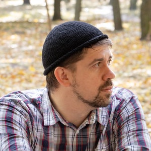 Può includere: Un uomo con un berretto nero e una camicia a quadri con le braccia incrociate, in piedi davanti a un parapetto di legno. Lo sfondo è un'immagine sfocata di alberi e foglie cadute.