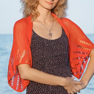 K&ouml;nnte beinhalten: Ein leuchtend orangefarbener Strick-Bolero mit einem durchbrochenen Muster und vertikalen Streifen aus &Ouml;sendetails. Das Kleidungsst&uuml;ck hat kurze, weite &Auml;rmel und eine drapierte Silhouette. Der Schriftzug ITS KNITS ist auf der linken Seite zu sehen.