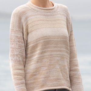 May include: A beige and tan striped sweater with long sleeves. The sweater has a round neckline and a relaxed fit. The sleeves have a subtle color variation. The person is wearing blue jeans. The background is a blurred view of water and sky.
