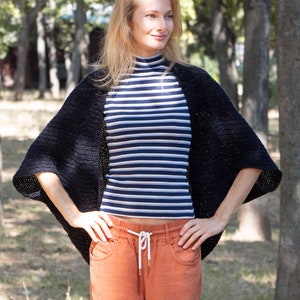 Könnte beinhalten: Eine Frau trägt einen schwarzen Strickcardigan über einem gestreiften Shirt und einer orangefarbenen Hose. Der Cardigan hat einen weiten, offenen Ausschnitt und lange Ärmel.