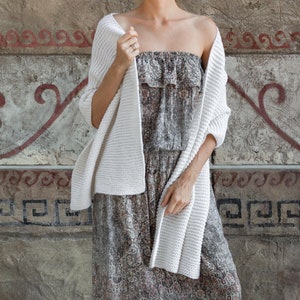 Peut inclure: Un cardigan blanc en tricot à la coupe ample et au motif texturé. Le cardigan est drapé sur les épaules d'une personne et porté avec une robe à motifs marrons et blancs.