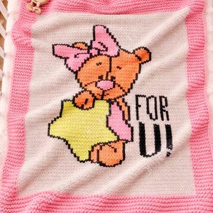 May include: A knitted baby blanket featuring a cartoon bear holding a star. The bear is orange and pink, with a pink bow. The blanket has a pink border and the words "FOR U" are printed on the blanket.